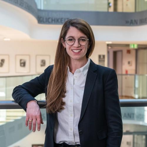 A smiling woman with long brown hair, wearing glasses, a white button-down shirt, and a dark blazer, stands leaning on a railing in a modern building atrium with abstract art and text on the walls in the background.